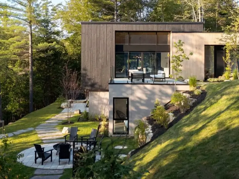 hillside house design with a wood-clad modern home and stucco lower level built into a green slope with a stone path and fire-pit patio
